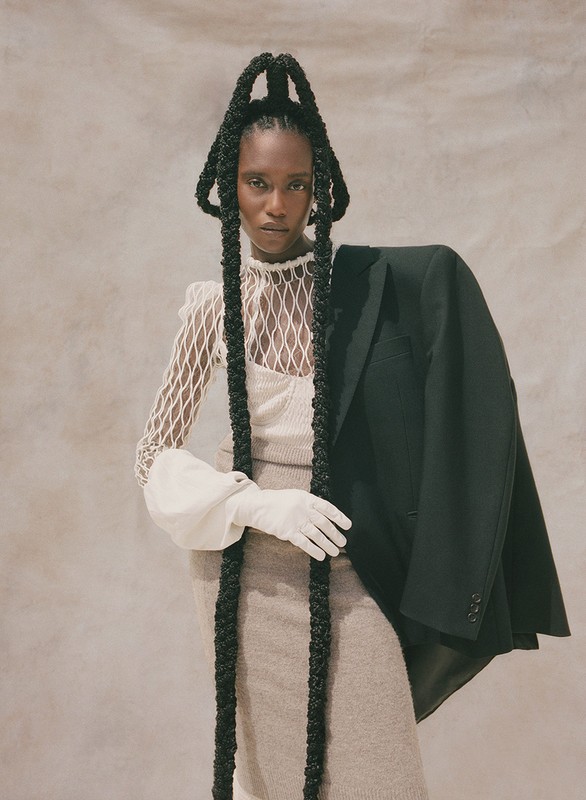 A Black woman poses indoors wearing a beige knit dress, a white fishnet top with puffy sleeves, a black jacket, and long braids.