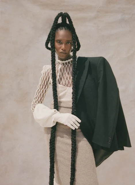 A Black woman poses indoors wearing a beige knit dress, a white fishnet top with puffy sleeves, a black jacket, and long braids.