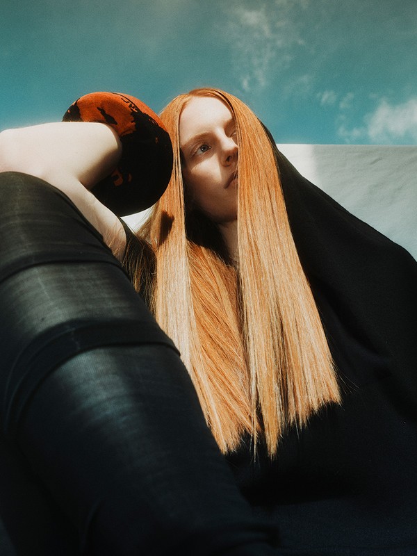 A red-haired woman with sleek hair, wearing black clothing, poses against a backdrop of a light blue sky with wispy white clouds.