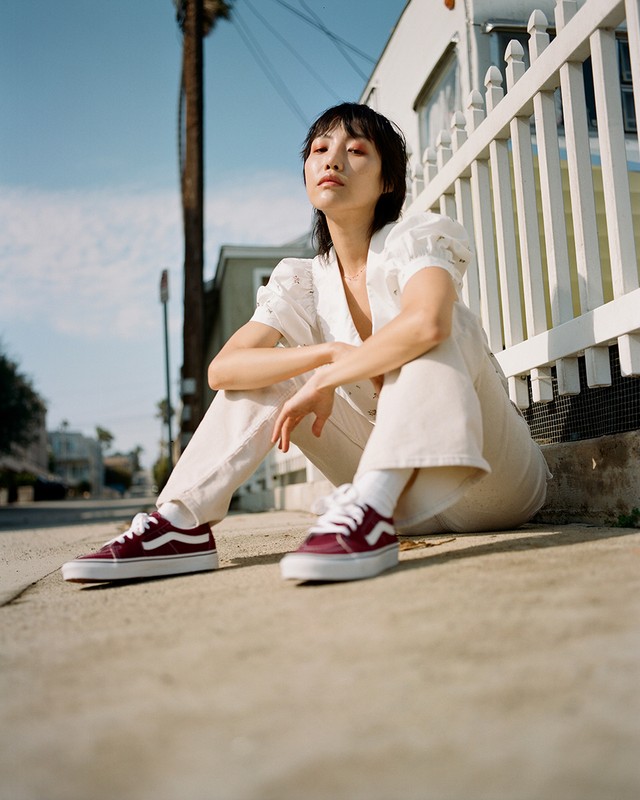 Woman sitting on sidewalk in white blouse and pants, wearing red Vans sneakers in an outdoor, daytime setting with trees and houses.