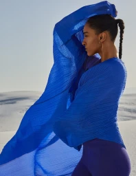 Woman in blue pleated top with fabric caught in the wind against a pale sandy desert landscape under a clear sky.