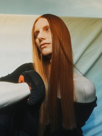 Portrait of a red-haired woman with long straight hair wearing a black top against a fabric backdrop by Phoebe Seligman