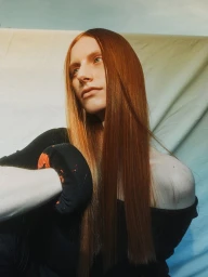 Portrait of a red-haired woman with long straight hair wearing a black top against a fabric backdrop by Phoebe Seligman