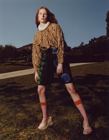 Red-haired model in zebra print blouse and patterned shorts standing on a grassy lawn with trees and a house in the background