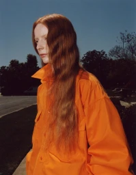 Red-haired woman wearing a bright orange collared shirt outdoors against a clear blue sky by Phoebe Seligman