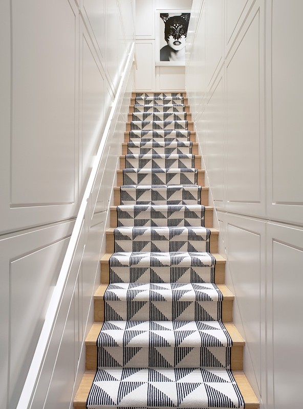 Hallway staircase with geometric patterned runner and white walls leading up to a framed artwork of a masked woman.