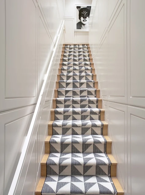 Hallway staircase with geometric patterned runner and white walls leading up to a framed artwork of a masked woman.