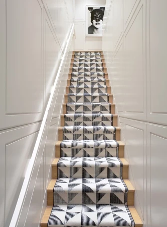 Hallway staircase with geometric patterned runner and white walls leading up to a framed artwork of a masked woman.