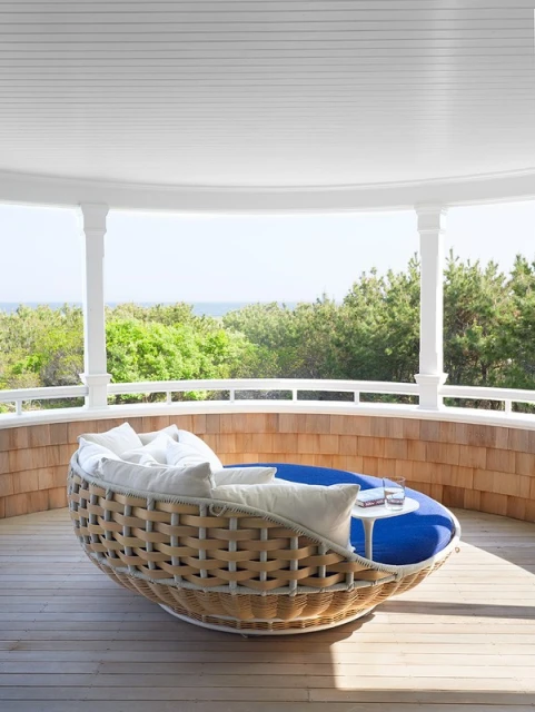 Wicker outdoor daybed with white cushions sits on a wooden porch with a cedar shingle wall and a view of greenery and the ocean.