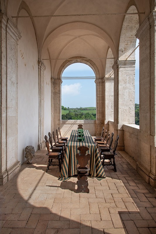 Stone archway frames an outdoor dining table set with striped tablecloth and green dishes, positioned on a stone patio with a rural view.