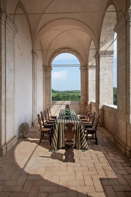 Stone archway frames an outdoor dining table set with striped tablecloth and green dishes, positioned on a stone patio with a rural view.