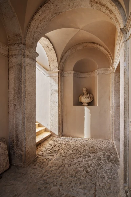 Interior hallway of old building with stone arches, stone floor, bust in wall niche, and steps leading to another level bathed in sunlight.