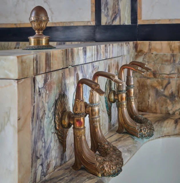 Close-up of three vintage brass water taps with swan neck spouts, mounted on a marble wall and surface, suggesting a luxurious bathroom.