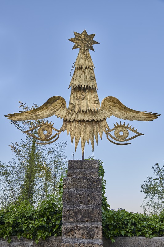 Golden sculpture of a tree with wings and eyes mounted on a weathered stone pedestal in an outdoor setting with blue sky background.