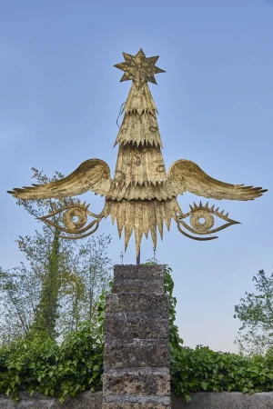 Golden sculpture of a tree with wings and eyes mounted on a weathered stone pedestal in an outdoor setting with blue sky background.