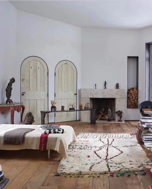 Interior of a living room featuring a daybed, textured rug, fireplace, art objects, white walls, and wooden floors in natural light.