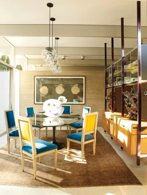 Contemporary dining room interior with round glass table, blue and yellow chairs, modern lighting, and display cabinet.
