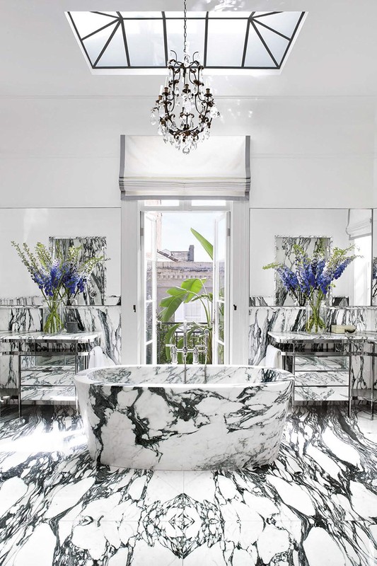 Luxurious bathroom interior featuring a marble bathtub, floor, and countertops, complemented by a chandelier and natural light from a window.