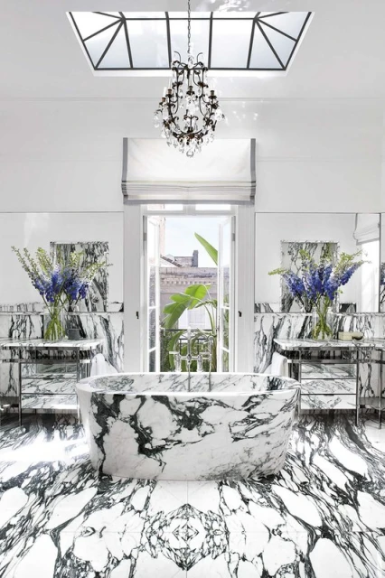 Luxurious bathroom interior featuring a marble bathtub, floor, and countertops, complemented by a chandelier and natural light from a window.