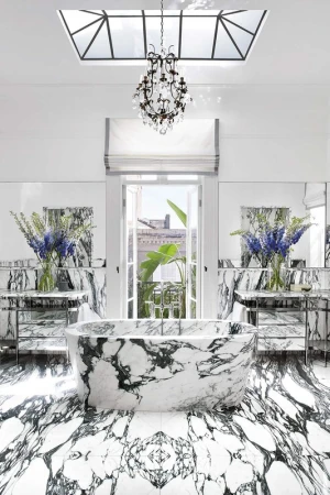 Luxurious bathroom interior featuring a marble bathtub, floor, and countertops, complemented by a chandelier and natural light from a window.