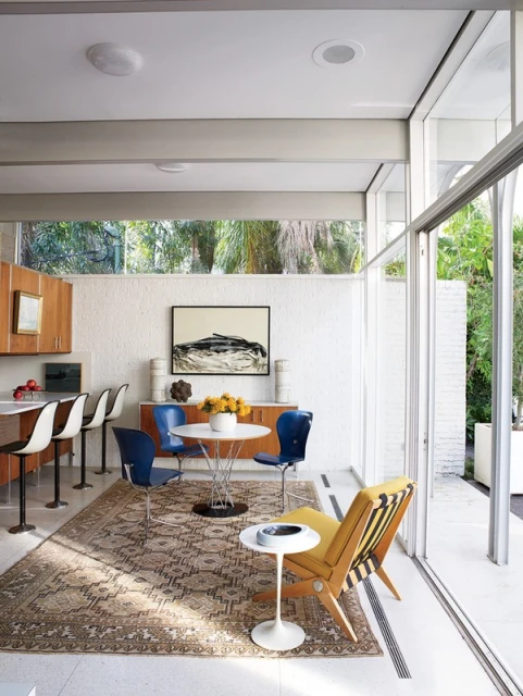 Mid-century modern dining room with a round table, blue chairs, and a yellow chair, complemented by neutral colors and ample natural light.