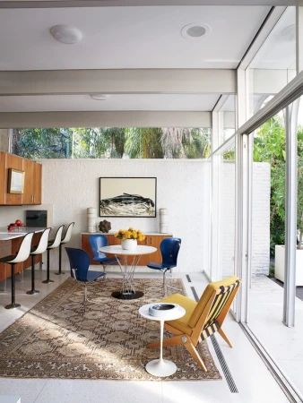 Mid-century modern dining room with a round table, blue chairs, and a yellow chair, complemented by neutral colors and ample natural light.