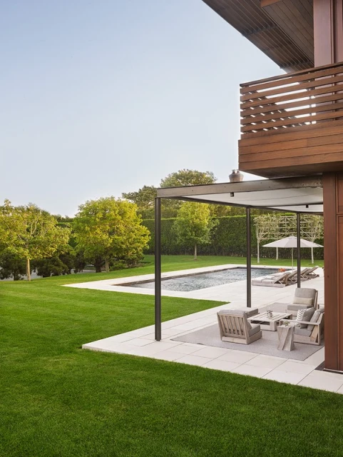 Exterior view of a modern house with a swimming pool, outdoor seating, a manicured lawn, and mature trees creating a serene backyard space.