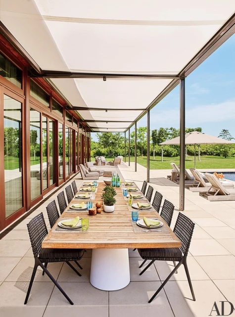 Outdoor dining area features a long wooden table with place settings, surrounded by black chairs on a stone patio with lounge seating.