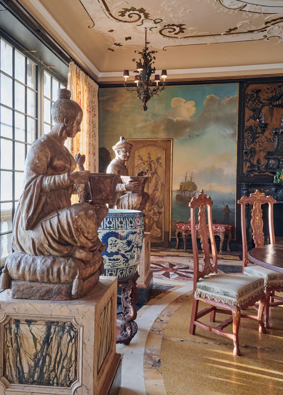 Ornate room with two sculptures of figures in traditional clothing, antique furniture, and a landscape mural wall decoration.