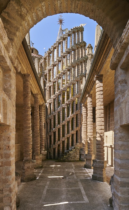 View through an archway revealing an intricate stone structure with a decorative spire, set against a clear blue sky and illuminated by sunlight.