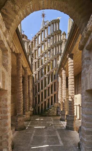 View through an archway revealing an intricate stone structure with a decorative spire, set against a clear blue sky and illuminated by sunlight.