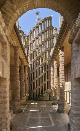 View through an archway revealing an intricate stone structure with a decorative spire, set against a clear blue sky and illuminated by sunlight.