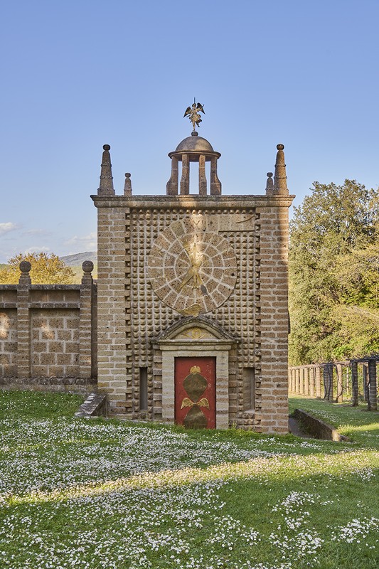 Ornate stone structure in a garden with a red door and green lawn with white flowers on a sunny day, architectural detail.