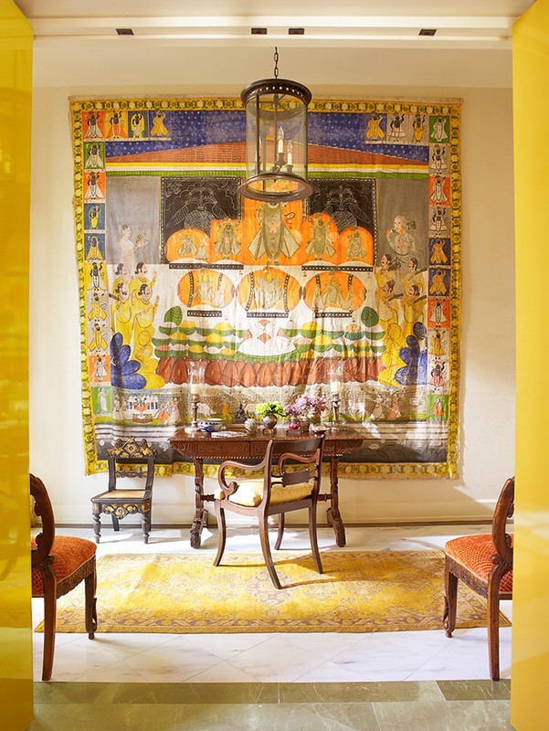 Interior view of a room with a colorful art tapestry hanging on the wall above a dark wood table and chairs, and a yellow rug.
