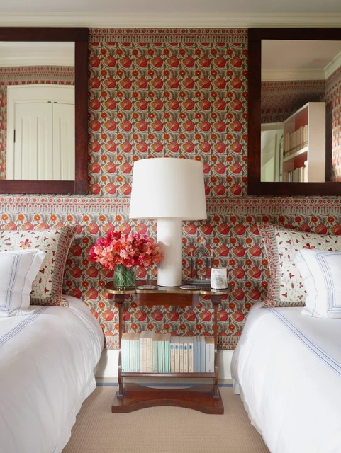 Symmetrical interior with two beds flanking a floral-wallpapered wall, centered by a lamp, flowers, and small books on a wooden table.