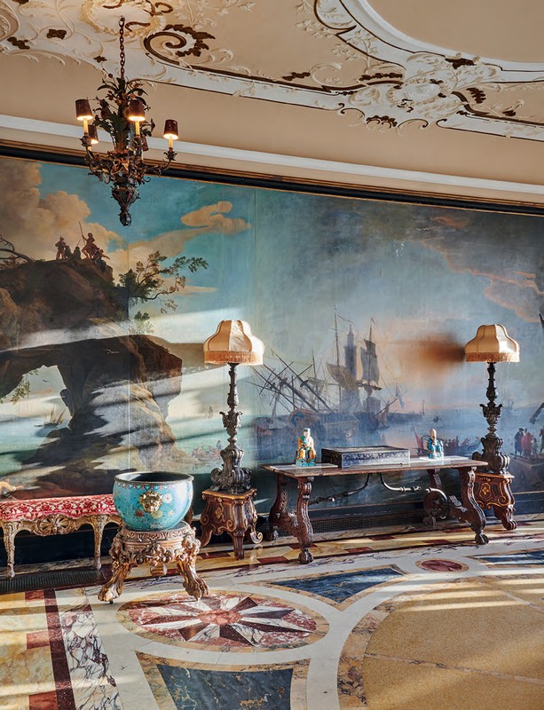 Ornate room with mural of seascape, antique furniture, chandelier, and patterned marble floor, illuminated by soft, diffused light.