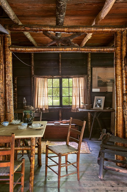Rustic interior scene with wooden furniture: table set with teacups, wood chairs, beams and posts, framed art, and a window.