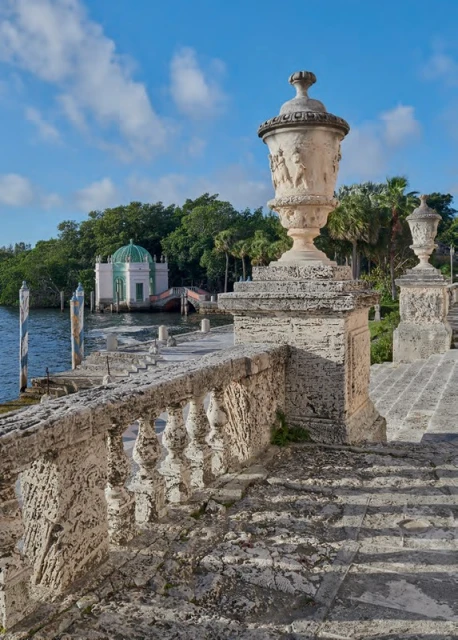 Stone staircase with decorative balustrade and urns leads to a gazebo on the water under a blue sky with light clouds.