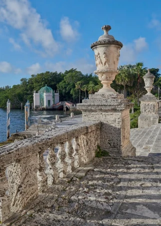 Stone staircase with decorative balustrade and urns leads to a gazebo on the water under a blue sky with light clouds.