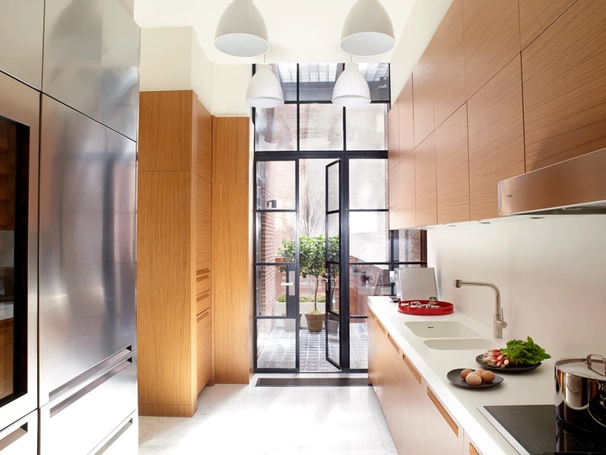 Modern kitchen with wood cabinets, stainless steel appliances, white countertops, and natural light through a large window with a courtyard view.