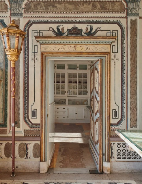 Interior shot of a vintage room with an open doorway, ornate decorative painting, a gold lantern, and a glimpse into a storage room.