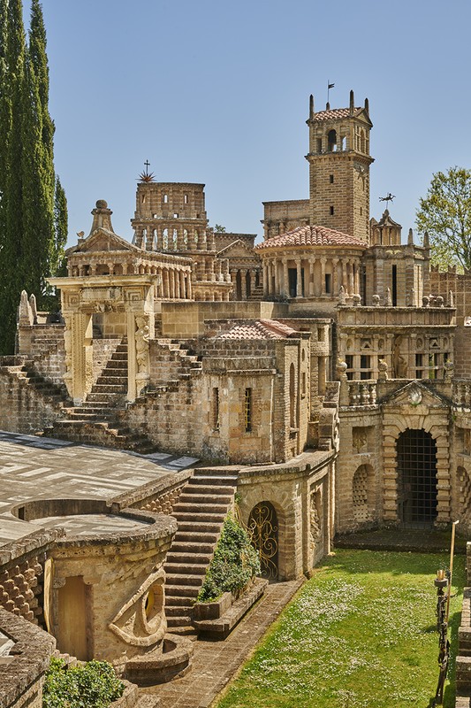 Complex architectural landscape featuring an Italianate tower and multiple stone structures with stairs and landscaped grass in bright daylight.