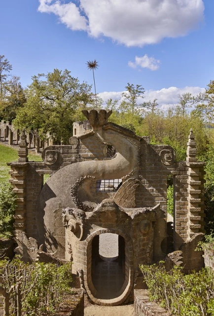 Stone dragon sculpture in Bomarzo Gardens, Italy, featuring weathered stone, lush greenery, and clear blue sky, showcasing Renaissance art.