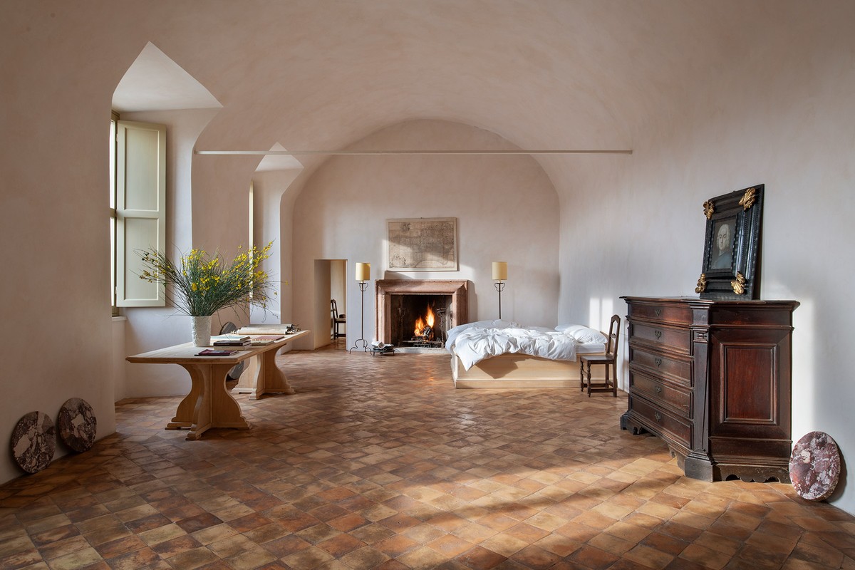 Bedroom interior featuring a large bed, fireplace, antique dresser, and terracotta tile flooring, bathed in natural light from arched windows.
