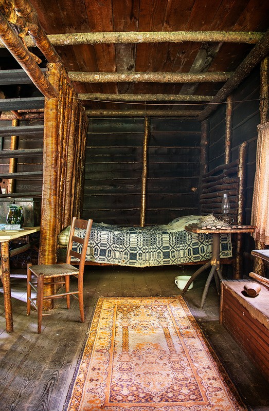 Rustic log cabin interior with a patterned rug, wooden chair, and a bed featuring a traditional quilt; cozy and vintage ambiance.