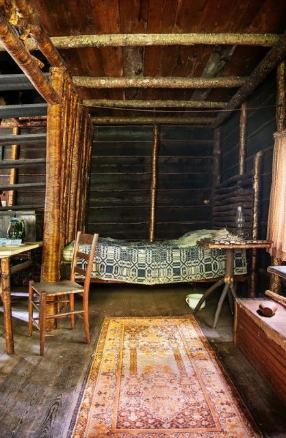 Rustic log cabin interior with a patterned rug, wooden chair, and a bed featuring a traditional quilt; cozy and vintage ambiance.