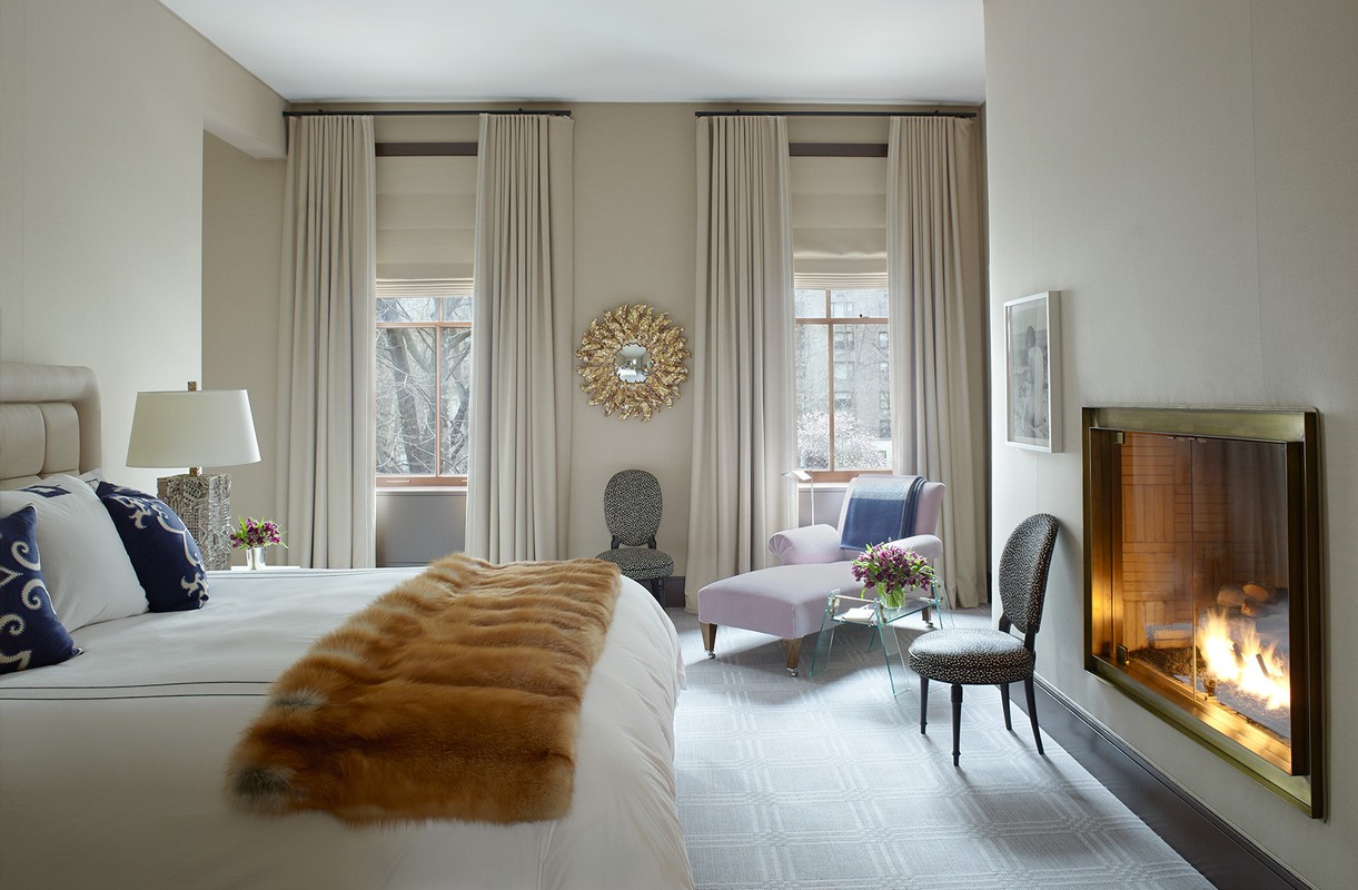 Bedroom interior featuring a bed with fur throw, chaise lounge, chairs, fireplace, windows with natural light, and neutral color palette.
