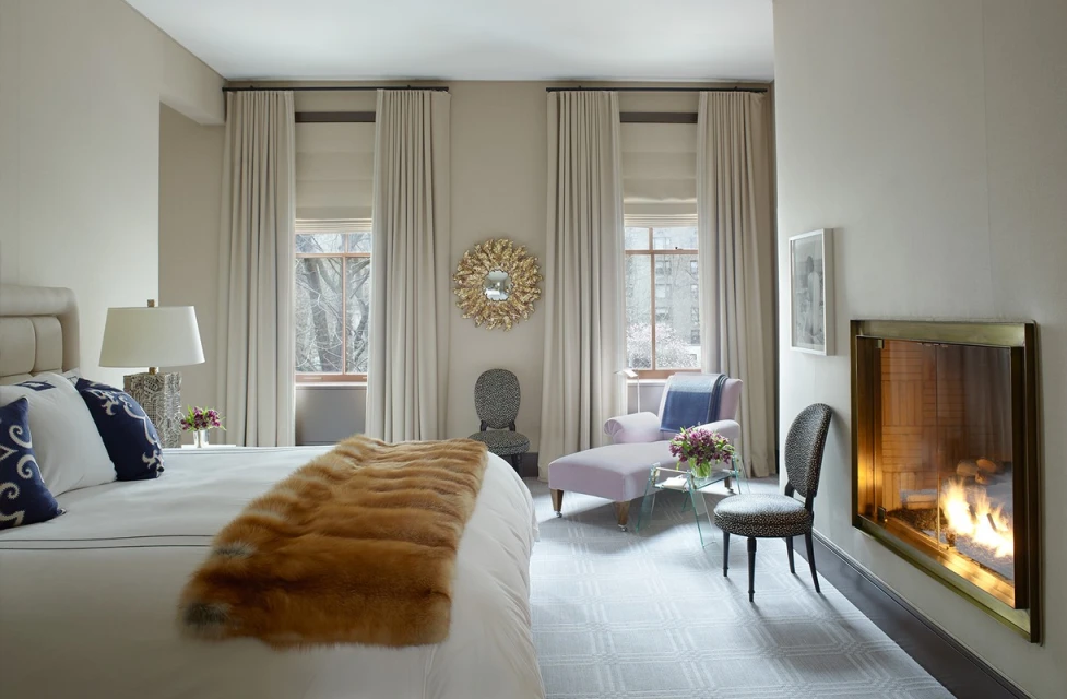 Bedroom interior featuring a bed with fur throw, chaise lounge, chairs, fireplace, windows with natural light, and neutral color palette.