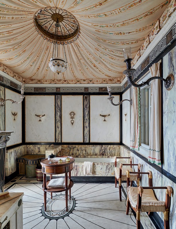 Ornate bathroom interior featuring marble walls and floor, a round wooden table, upholstered chairs, and decorative lighting fixtures.