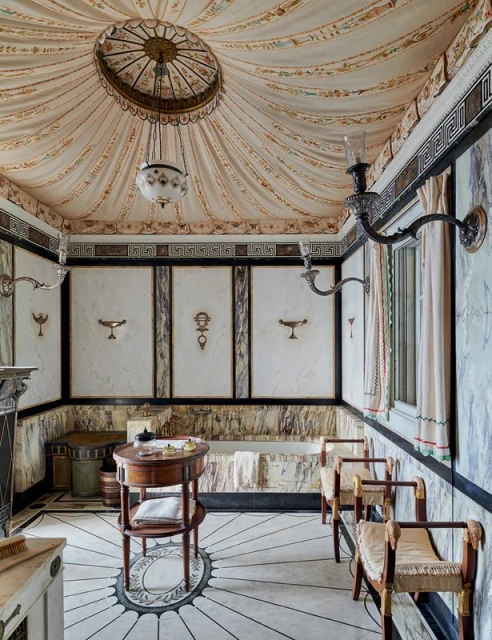 Ornate bathroom interior featuring marble walls and floor, a round wooden table, upholstered chairs, and decorative lighting fixtures.